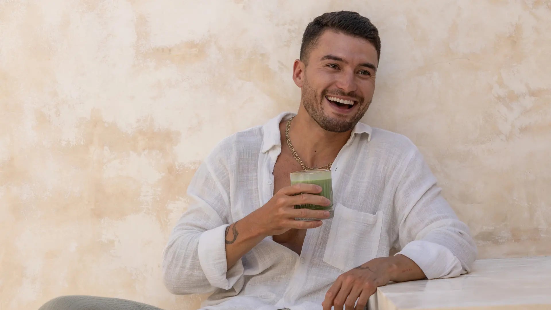 Smiling man in a white shirt enjoying a glass of moringa latte while sitting against a textured beige wall.