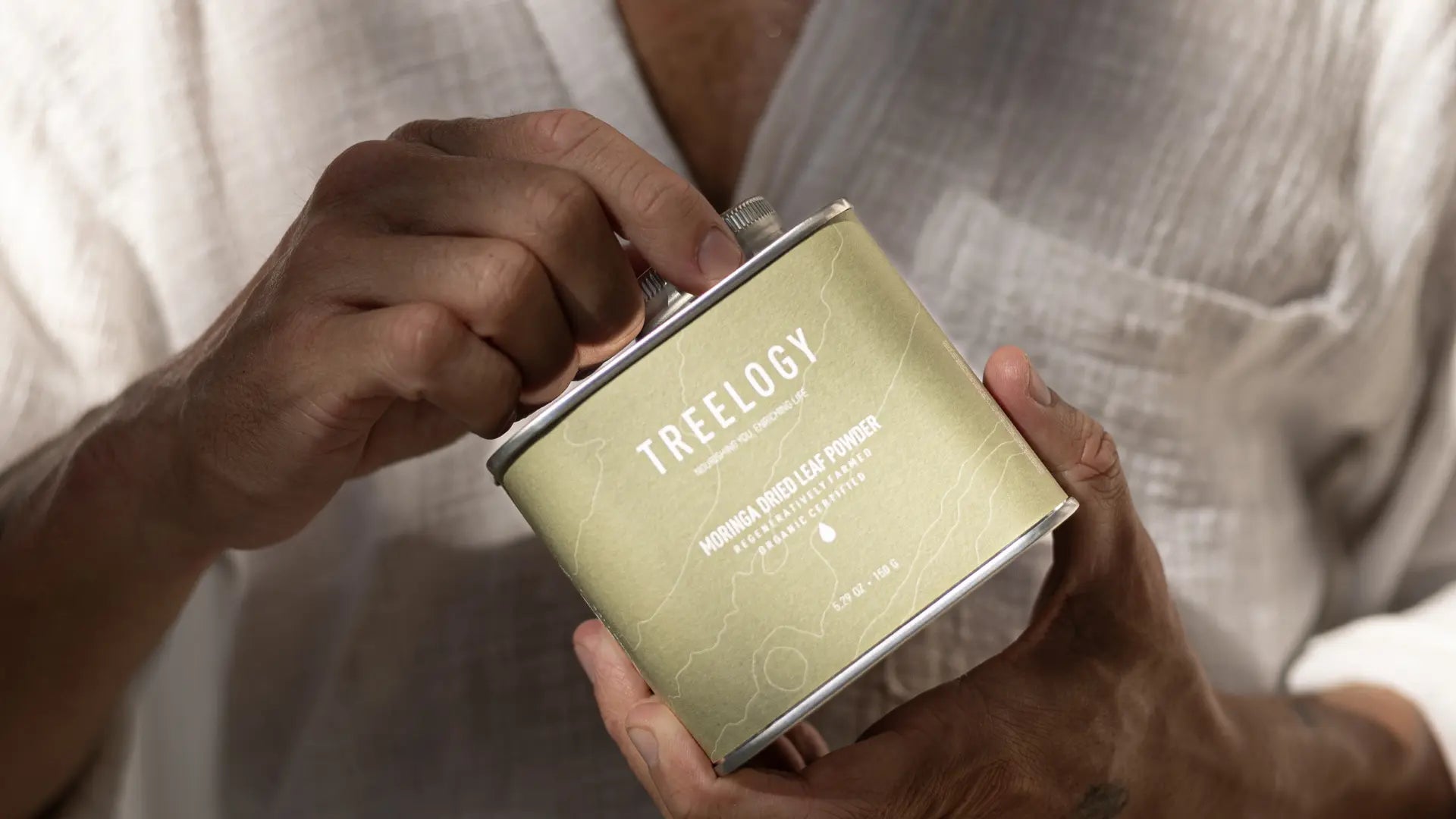 Close-up of hands opening a Treelogy moringa dried leaf powder tin, with a person wearing a light textured shirt in the background.