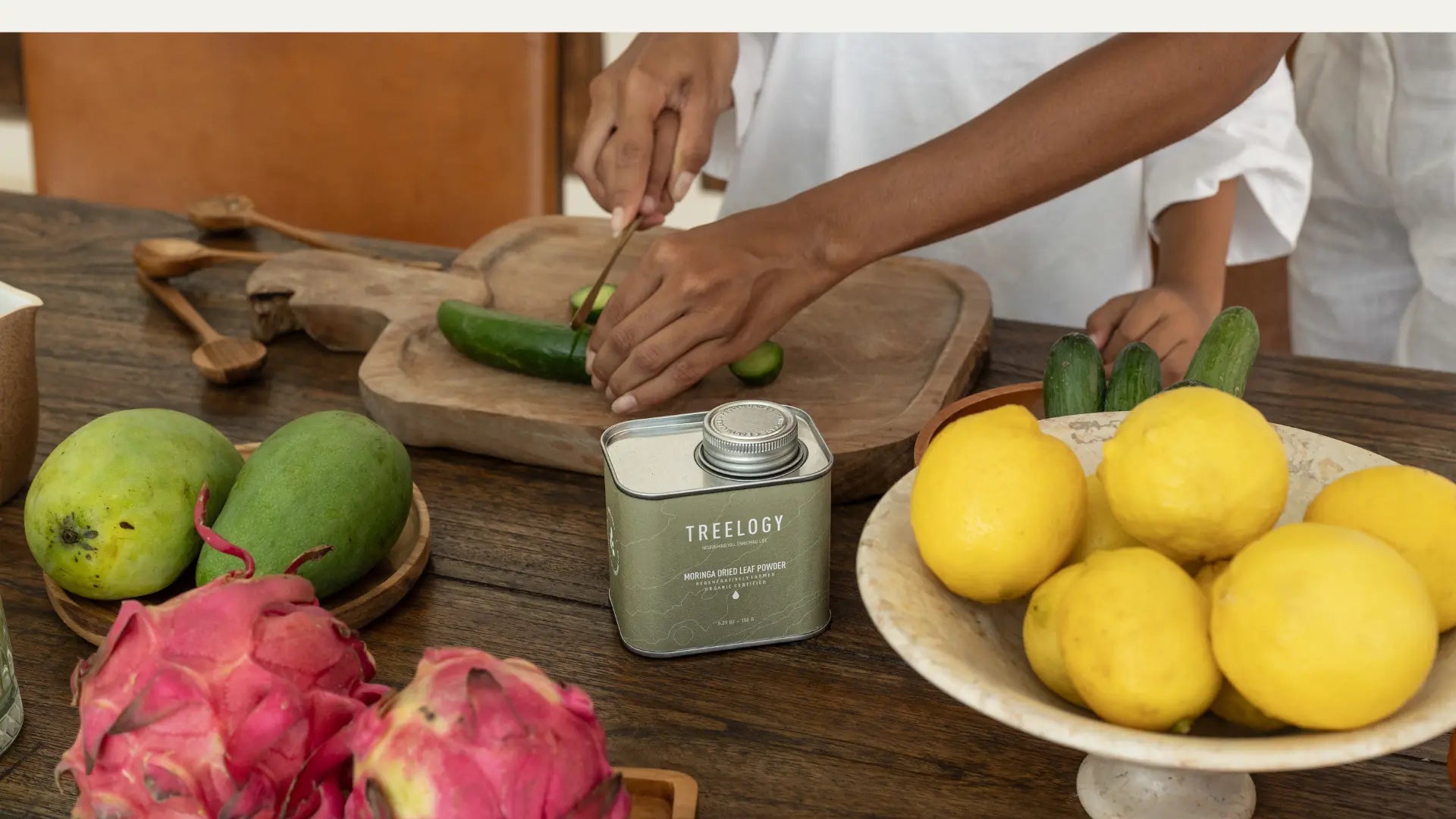 Hands slicing cucumber on a wooden board surrounded by fresh fruits and a tin of Treelogy Moringa Dried Leaf Powder.