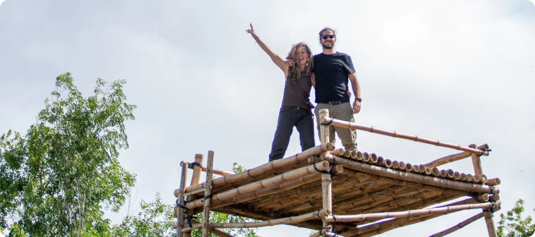 Two people standing on top of a bamboo structure surrounded by trees, with one raising an arm in the air.
